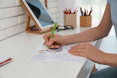 Young woman coloring antistress page at desk indoors, closeup
