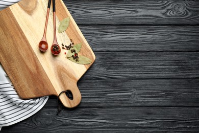 Cutting board and condiments on black wooden table, flat lay with space for text. Cooking utensils