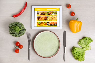 Modern tablet with open page for online food ordering, scattered vegetables, plate and cutlery on white wooden table, flat lay. Concept of delivery service