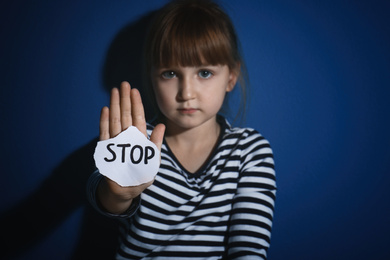 Abused little girl with sign STOP near blue wall, focus on hand. Domestic violence concept
