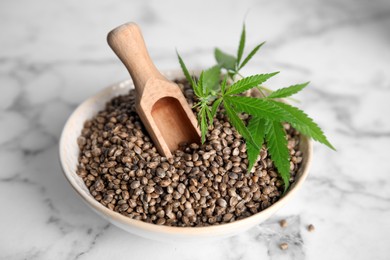 Bowl with hemp seeds, wooden scoop and fresh leaves on white marble table, closeup