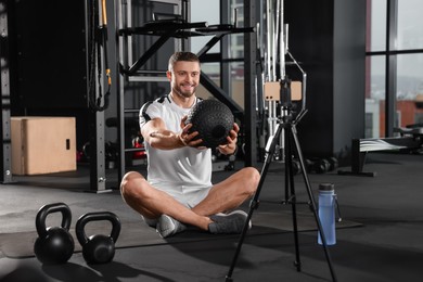 Photo of Man with ball streaming online training on phone at gym. Fitness coach