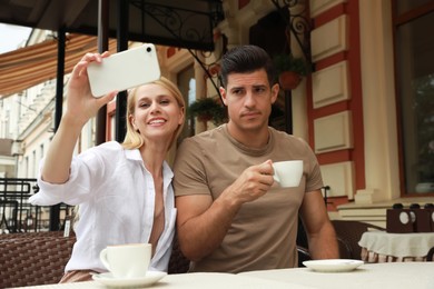Young woman trying to take selfie with her displeased boyfriend in outdoor cafe. Boring date