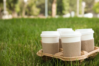 Takeaway paper coffee cups with plastic lids and sleeves in cardboard holder on green grass outdoors, space for text