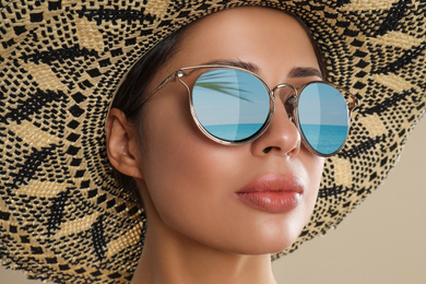 Beautiful young woman wearing sunglasses with reflection of sea and hat on beige background 