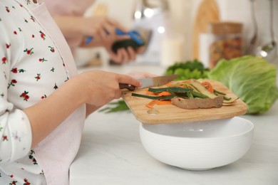 Little girl with cutting board and knife scraping vegetable peels into bowl on kitchen counter, closeup