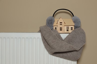 Modern radiator with earmuffs, scarf and wooden house near beige wall indoors, space for text. Winter heating efficiency
