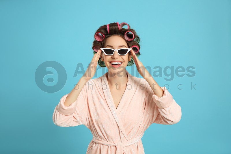 Beautiful young woman in bathrobe with hair curlers and sunglasses on light blue background