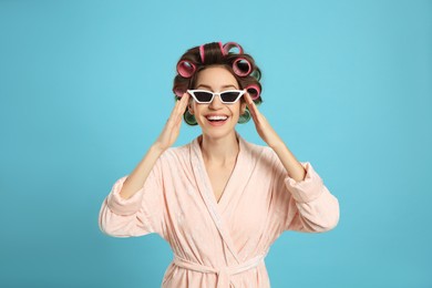 Beautiful young woman in bathrobe with hair curlers and sunglasses on light blue background