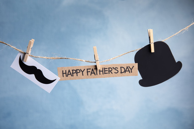 Photo of Card with words HAPPY FATHER'S DAY, paper mustache and hat hanging on rope against light blue background