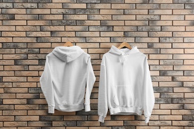Photo of New hoodie sweaters with hangers on brick wall. Mockup for design