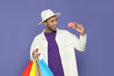 Photo of Happy African American man in hat with shopping bags and credit card on purple background