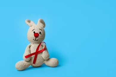 Cute knitted toy bunny with red ribbon on blue background, space for text. AIDS disease awareness
