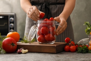 Woman putting tomatoes into glass jar at grey kitchen table, closeup. Pickling vegetables