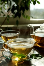 Fresh green tea in glass cups with saucers, leaves and teapot on wooden table