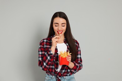 Beautiful young woman eating French fries on grey background