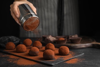 Photo of Woman covering delicious chocolate truffles with cocoa powder at black table, closeup