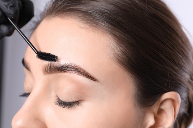 Beautician brushing woman's eyebrows after tinting on light background, closeup