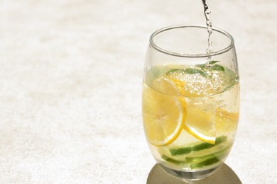 Pouring water into glass with lemon slices and mint at light grey table, closeup. Space for text