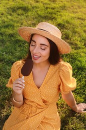 Beautiful young woman eating ice cream glazed in chocolate on green grass outdoors