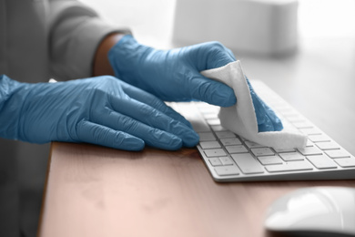 Woman in latex gloves cleaning computer keyboard with wet wipe at table, closeup