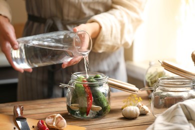 Woman pouring water into jar with cucumbers at wooden table, closeup. Pickling vegetables