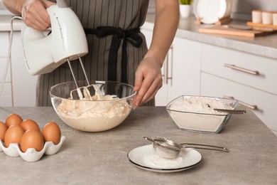 Woman whipping white cream with mixer at light grey table in kitchen, closeup