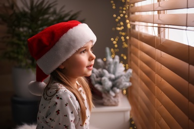 Photo of Cute little girl in Christmas hat near window at home