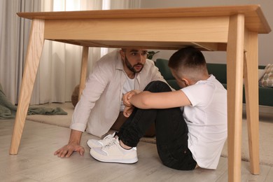 Father comforting his scared son under table in living room during earthquake