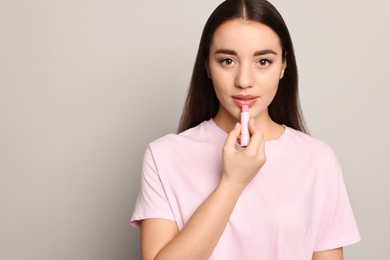 Young woman applying lip balm on grey background, space for text