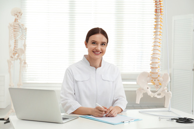 Female orthopedist with laptop near human spine model in office