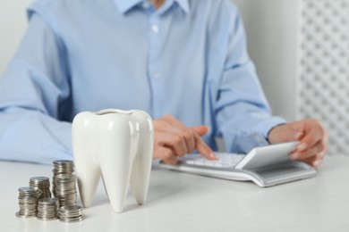 Woman using calculator at white table indoors, focus on ceramic model of tooth and coins. Expensive treatment