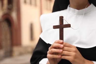 Young nun holding Christian cross near building outdoors, closeup. Space for text