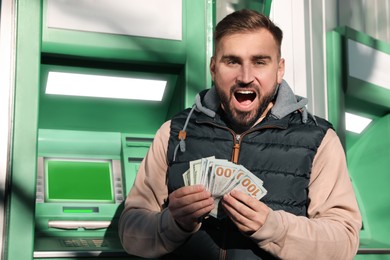 Excited young man with money near cash machine outdoors