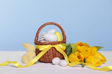 Photo of Wicker basket with festively decorated Easter eggs, bunny and beautiful tulips on white wooden table against light blue background
