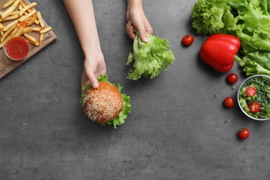 Top view of woman choosing between vegetables and burger with French fries at black table, closeup