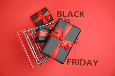 Small shopping cart with gift boxes and phrase Black Friday on red background, top view