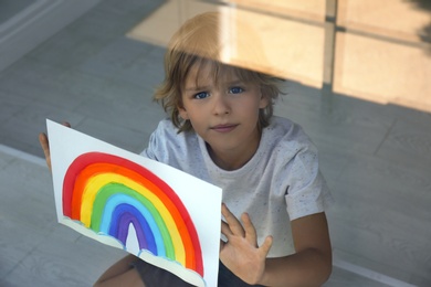 Little boy with picture of rainbow near window, view from outdoors. Stay at home concept