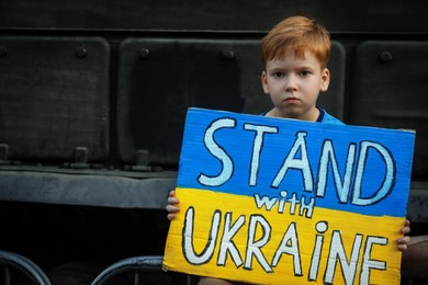 Sad boy holding poster in colors of national flag and words Stand With Ukraine near broken military tank outdoors. Space for text