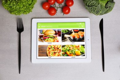Modern tablet with open page for online food ordering, vegetables and cutlery on grey table, flat lay. Concept of delivery service