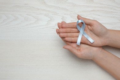 Woman holding light blue awareness ribbon at white wooden table, top view. Space for text