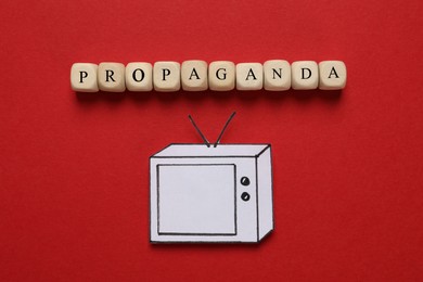 Photo of Word Propaganda made of wooden cubes and paper TV on red background, flat lay. Information warfare concept