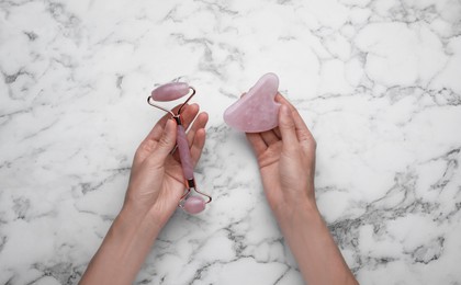 Photo of Woman holding gua sha stone and face roller at white marble table, top view