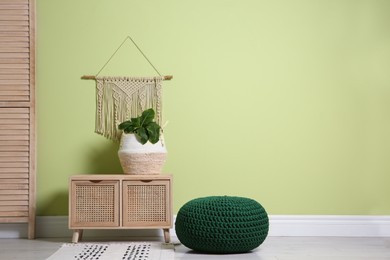 Stylish room interior with comfortable knitted pouf, wooden furniture and plant near light green wall, space for text