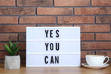 Lightbox with phrase Yes You Can, cup of coffee and potted houseplant on table against brick wall. Motivational quote
