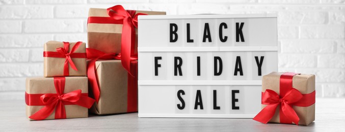 Lightbox with words Black Friday Sale and gift boxes on wooden table against white brick wall. Banner design