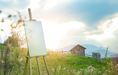 Wooden easel with blank canvas and picturesque view of village on mountain slopes