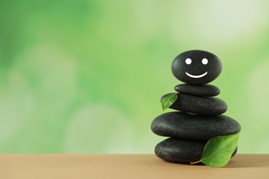 Stack of stones with drawn happy face and green leaves on table against blurred background, space for text. Zen concept