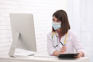 Photo of Pediatrician in protective mask consulting patient online at table indoors
