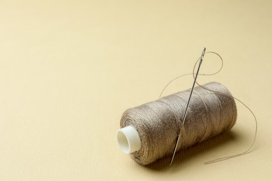 Spool of grey sewing thread with needle and thimble on beige background. Space for text